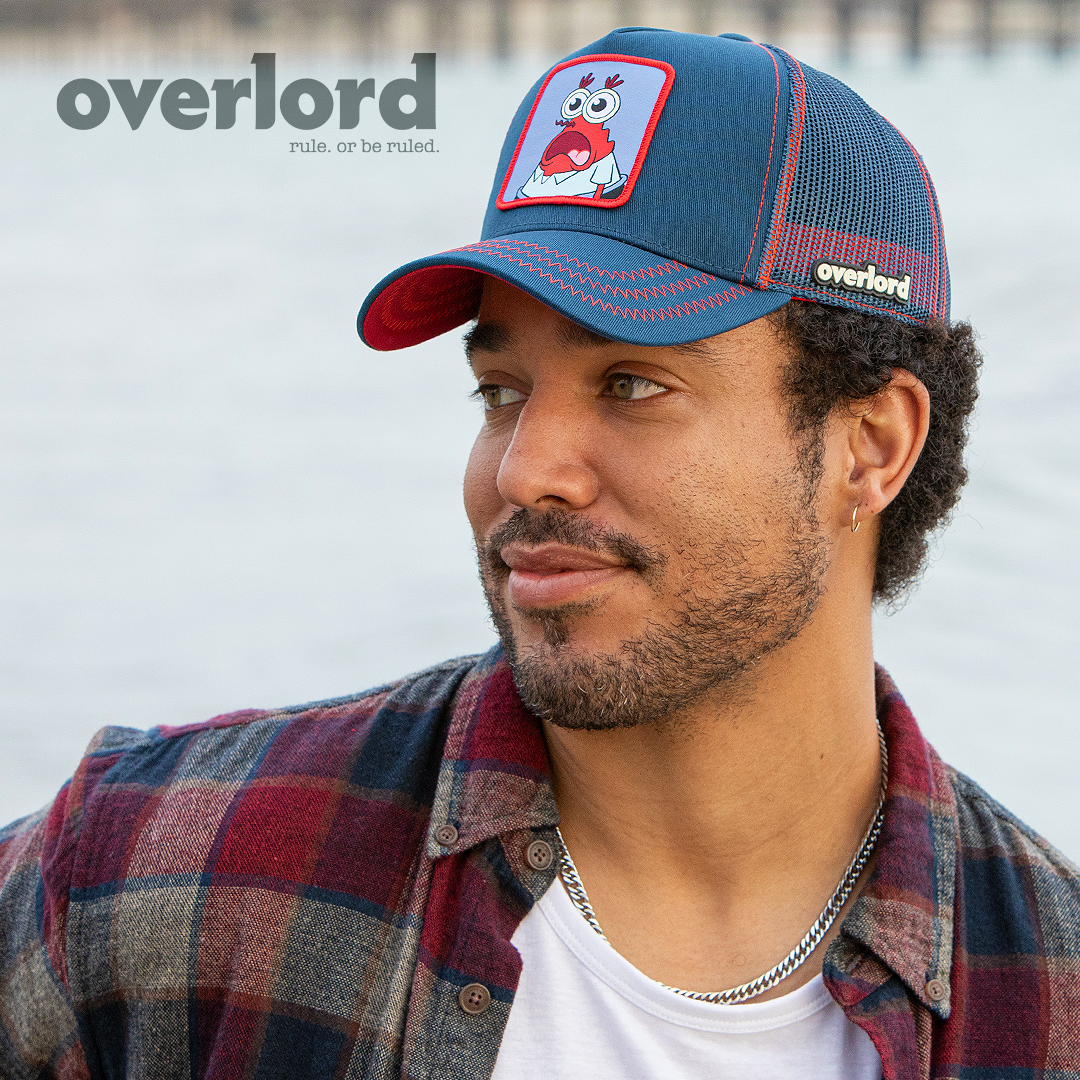 Man wearing navy OVERLORD X SpongeBob Krusty Krab surprised face trucker baseball cap with red zig zag stitching. PVC Overlord logo.