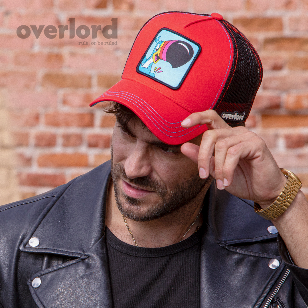 Man wearing red OVERLORD X Kelloggs Toucan Sam Froot Loops trucker baseball cap hat with blue stitching. PVC Overlord logo.