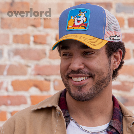 Man wearing blue and orange OVERLORD X Kelloggs Tony Tiger Frosted Flakes trucker baseball cap hat with black zig zag stitching. PVC Overlord logo.