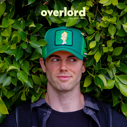 Man in bush wearing green OVERLORD X The Simpsons present Homer in bush trucker baseball cap hat with green stitching. PVC Overlord logo.