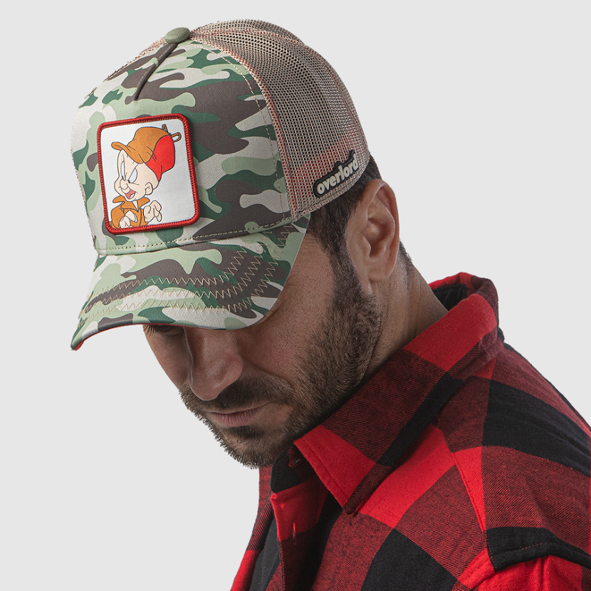 Man wearing camo OVERLORD X Looney Tunes Elmer Fudd trucker baseball cap hat with khaki zig zag stitching. PVC Overlord logo.