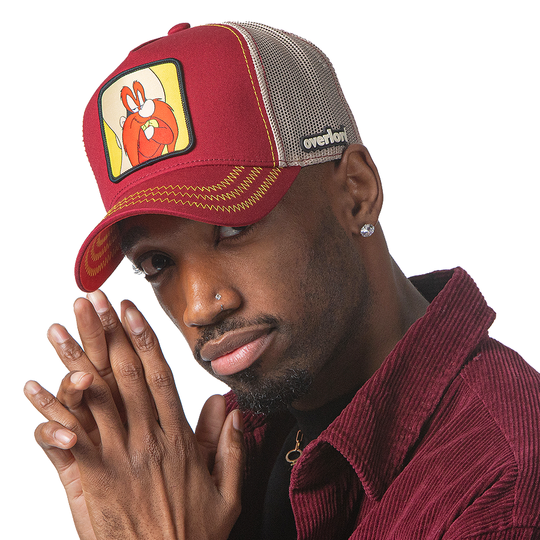 Man wearing dark red OVERLORD X Looney Tunes smug Yosemite Sam trucker baseball cap hat with yellow zig zag stitching. PVC Overlord logo.