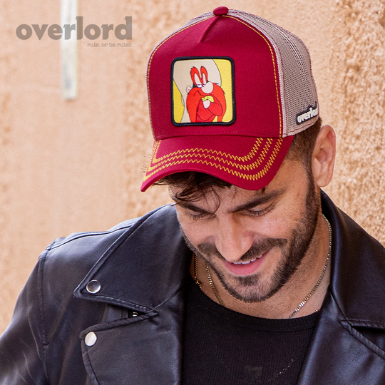 Man wearing dark red OVERLORD X Looney Tunes smug Yosemite Sam trucker baseball cap hat with yellow zig zag stitching. PVC Overlord logo.