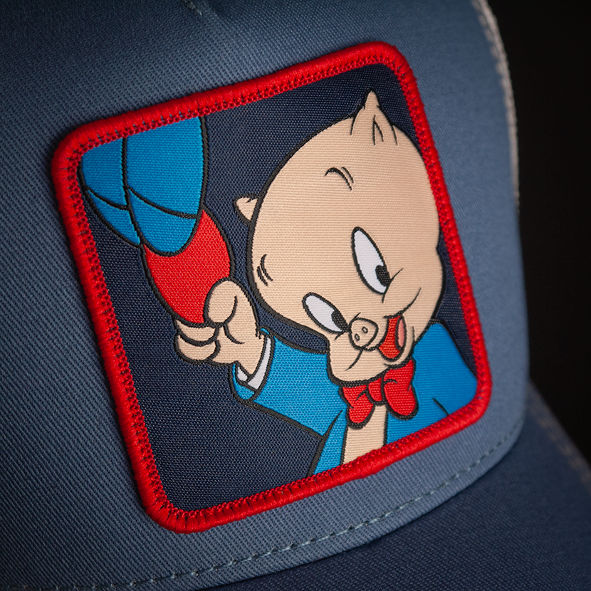 Blue OVERLORD X Looney Tunes Porky Pig holding a hat trucker baseball cap hat with woven Overlord patch closeup.