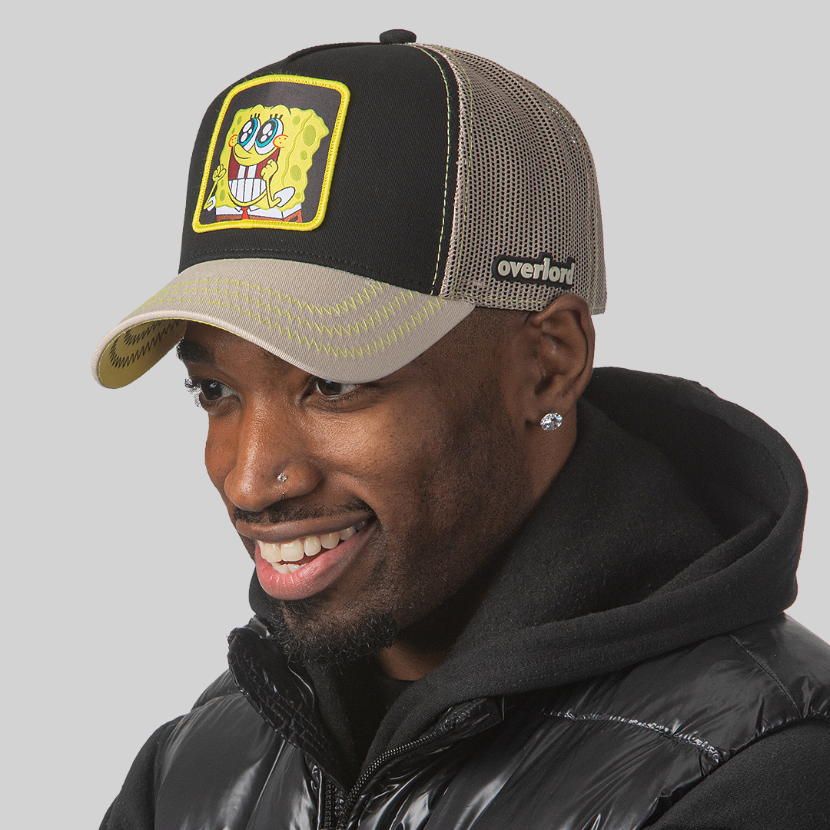 Man wearing black and tan OVERLORD X SpongeBob Big Smile SpongeBob trucker baseball cap hat with yellow zig zag stitching. PVC Overlord logo.