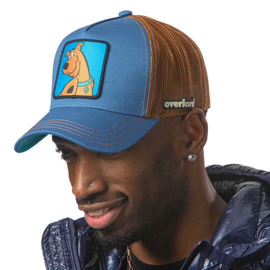 Man wearing blue OVERLORD X Scooby-Doo smiling Scooby trucker baseball cap hat with brown stitching. PVC Overlord logo.