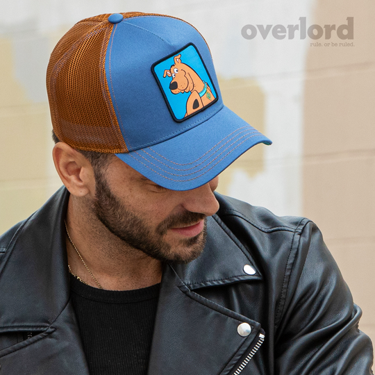 Man wearing blue OVERLORD X Scooby-Doo smiling Scooby trucker baseball cap hat with brown stitching. PVC Overlord logo.